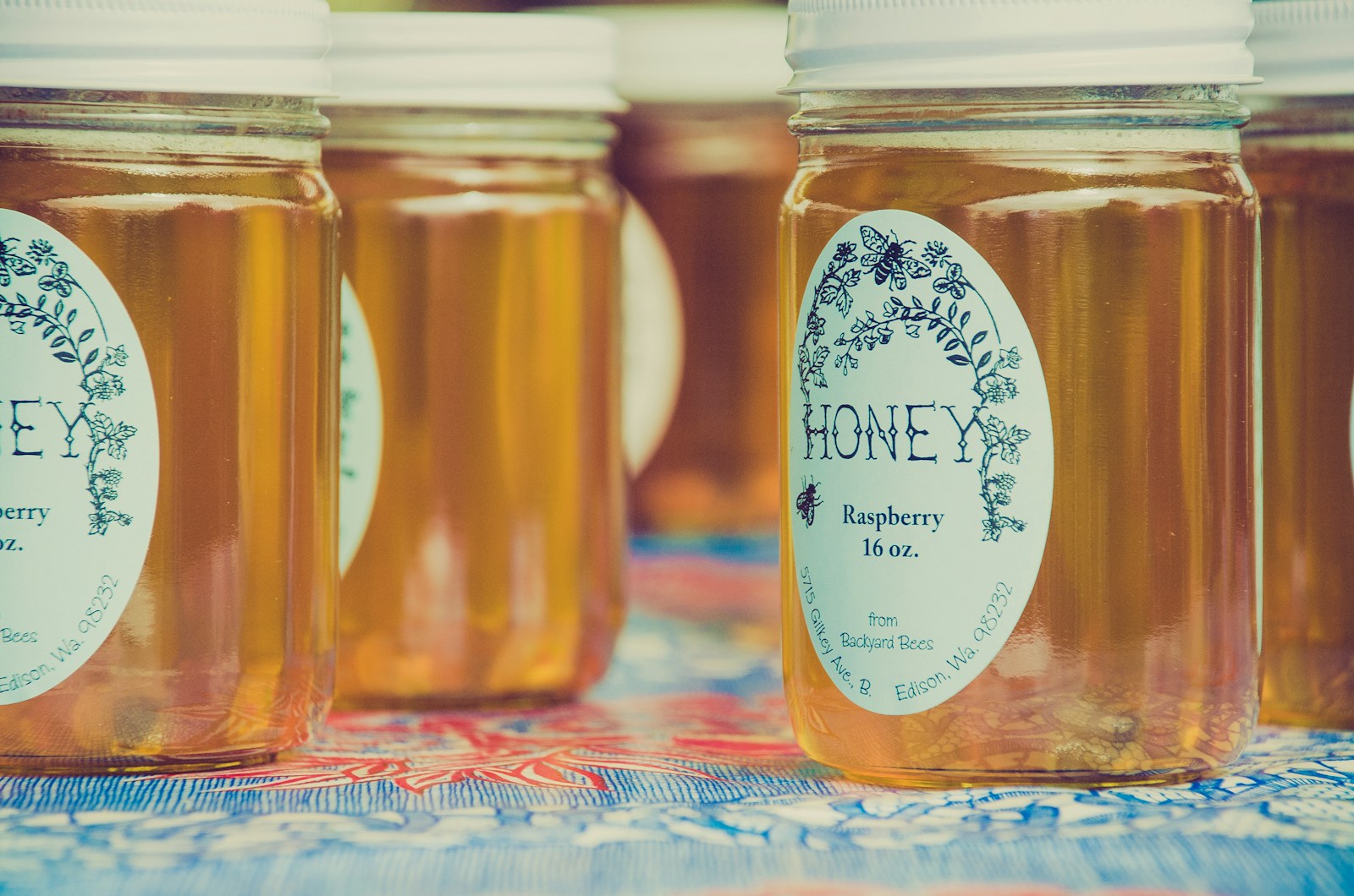 Słoiki z miodem malinowym o pojemności 16 oz, z etykietą przedstawiającą nazwę produktu i informacje o pochodzeniu z Backyard Bees, na kolorowym tle.