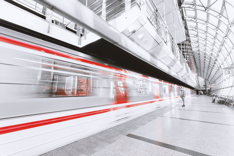 Stacja metra z nowoczesną architekturą, na której pociąg z szybkością przejeżdża przez peron. Widoczny jest także pasażer czekający na przyjazd transportu publicznego. Przestrzeń charakteryzuje się jasnym oświetleniem i minimalistycznym wystrojem.