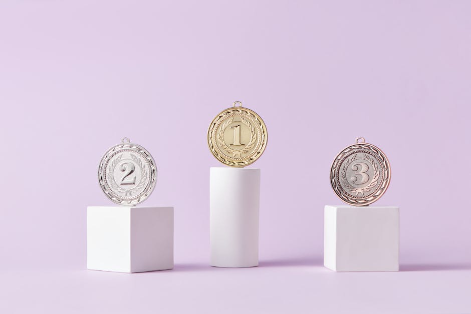 Medale sportowe na białych podium, symbolizujące osiągnięcia i rywalizację. Medal złoty, srebrny i brązowy umieszczone na trzech różnych postumentach, na tle pastelowej fioletowej ściany. Idealny obrazek do zilustrowania sukcesów, nagród i duchu sportowej rywalizacji.