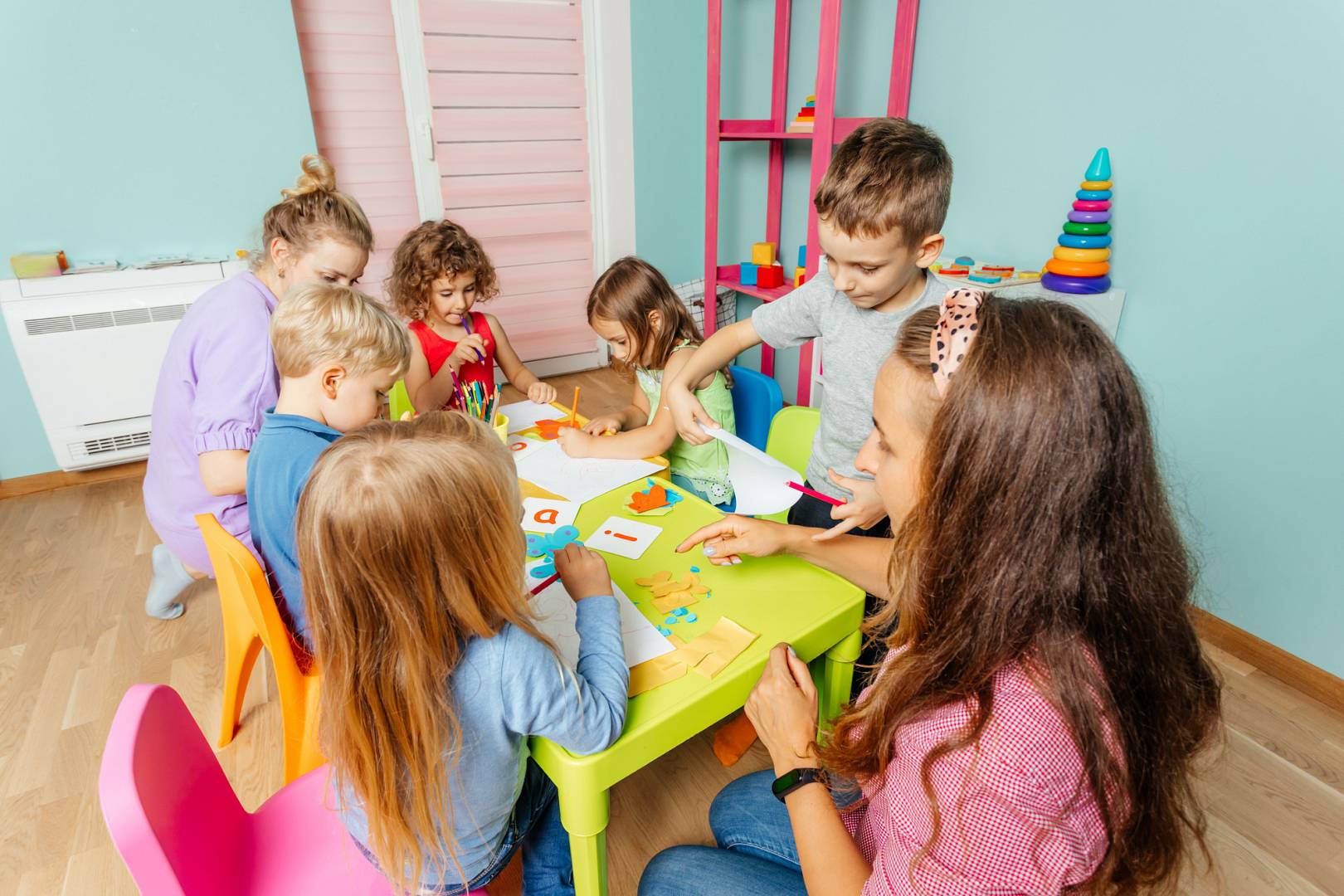 Grupa dzieci i nauczycielka bawią się przy kolorowym stole, tworząc prace plastyczne. W tle widoczne są kolorowe zabawki oraz przybory artystyczne.