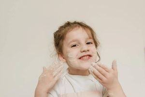 Dziecko z uśmiechem na twarzy, z kremem na rękach i twarzy, w białym ubraniu, na jasnym tle.